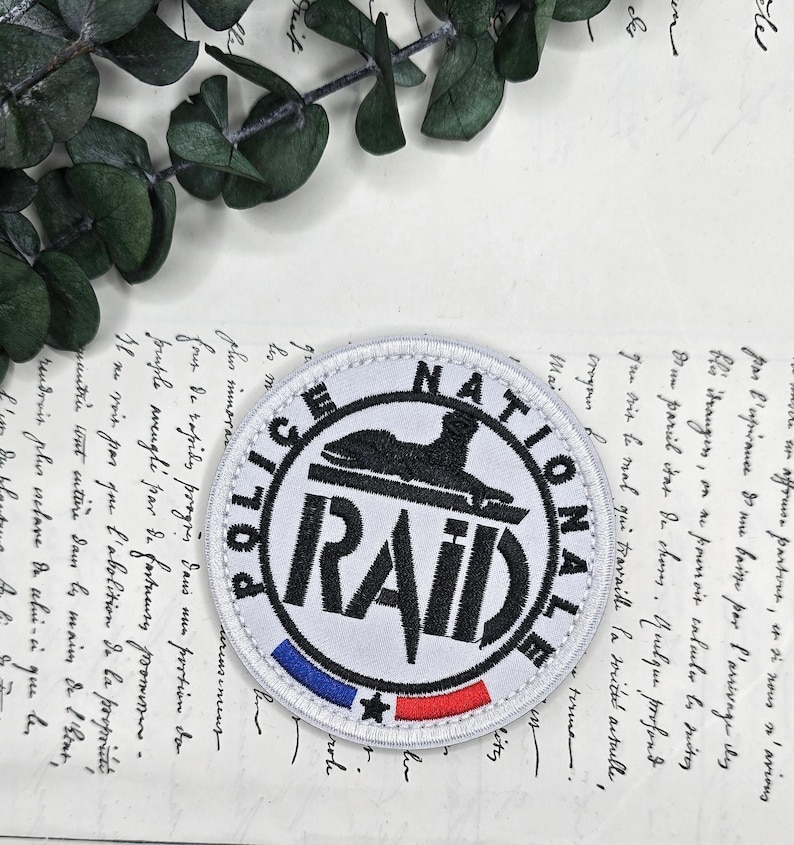 May include: A circular patch with the text "POLICE NATIONALE RAID". The patch is white with black text and a black emblem. It has a blue, white, and red stripe at the bottom. The patch is on a vintage paper background.