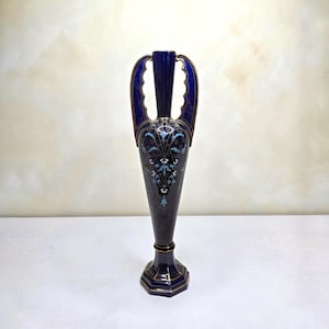May include: A tall, slender, dark blue ceramic vase with two ornate handles. The body features intricate white and light blue floral patterns with gold trim accents. It stands on a hexagonal base and has a narrow neck, showcasing a glossy finish.