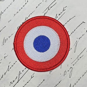 May include: Embroidered patch featuring a circular design with concentric rings of red, white, and blue. The patch has a red border, a white middle ring, and a blue center. The patch is likely a decorative emblem or insignia.