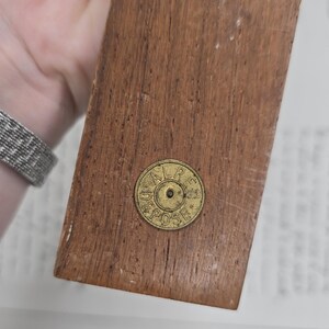 May include: A rectangular wooden object with a brass-colored circular emblem. The wood has a warm brown tone with visible grain. The emblem has the text "ALR DEPOSE" around the edge. The object is held by a person.