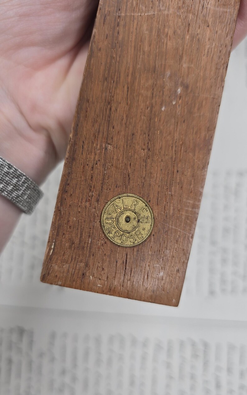 May include: A rectangular wooden object with a brass-colored circular emblem. The wood has a warm brown tone with visible grain. The emblem has the text "ALR DEPOSE" around the edge. The object is held by a person.