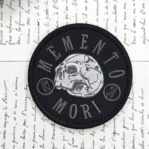 Puede incluir: Parche circular negro con las palabras "MEMENTO MORI" y un diseño de calavera, bordado con hilo gris. El parche es un accesorio oscuro, probablemente para ropa o bolsos.