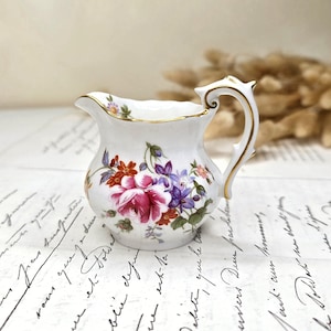 Könnte beinhalten: Ein kleines Milchkännchen aus weißem Porzellan mit einem bunten Blumenmuster aus rosa Rosen und violetten Glockenblumen. Das Kännchen hat einen goldfarbenen Rand und einen verzierten Henkel mit Goldakzenten, was ein filigranes Design ergibt.