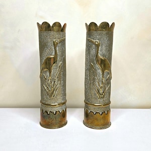 May include: A pair of tall, cylindrical brass vases featuring a textured surface with a raised relief design of a crane standing among reeds. The top and bottom edges of each vase have a scalloped, decorative border with a polished brass finish.
