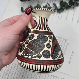 Peut inclure: Petit vase en céramique crème avec une anse, orné d'un motif de hibou noir et rouge. Le vase a un col étroit avec des rayures verticales noires et rouges et une bande rouge à la base. Le hibou est représenté avec des détails complexes.