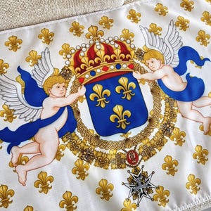 Op de afbeelding: Witte zijden sjaal met een gedetailleerd ontwerp met een koninklijk wapen, kroon en cherubs. Het wapen is blauw met gouden fleur-de-lis symbolen. Gouden fleur-de-lis patronen zijn verspreid over de witte achtergrond. De kroon is rood en goud.