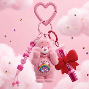 Pode incluir: Um porta-chaves rosa com fecho em forma de coração, uma figura Care Bear com um emblema de arco-íris na barriga, um pendente de contas com a palavra CHLOE e um tubo de brilho labial adornado com um grande laço vermelho. O urso é flocado.