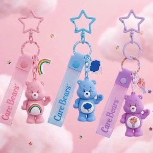 Może przedstawiać: Trzy breloki Care Bears z zapięciami w kształcie gwiazdek i paskami na nadgarstek. Różowy brelok przedstawia misia z tęczowym symbolem na brzuchu, niebieski misia z chmurką, a fioletowy misia z symbolem babeczki na brzuchu.