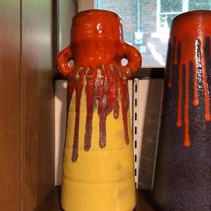 May include: A tall ceramic vase with a striking design. The top is a glossy orange, with two handles. The middle section is yellow, and the orange glaze drips down. A second vase is partially visible.