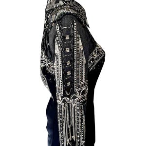 May include: A black denim jacket adorned with silver and clear embellishments. The jacket has a collared neckline and long sleeves, with detailed patterns of rhinestones and metallic accents. The design is heavily ornamented, creating a glamorous aesthetic.