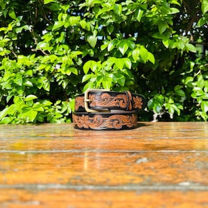 May include: A black leather belt featuring an intricate, hand-tooled floral pattern with natural tan highlights. The belt is equipped with a metallic brass-toned buckle and a matching leather keeper loop, showcasing detailed craftsmanship throughout.