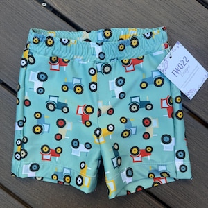 May include: Light blue toddler shorts with an all-over pattern of colorful tractors. The shorts have an elastic waistband and a tag that reads "TWO22 Designs". The tractors are in red, blue, yellow, and white.