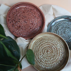 Peut inclure: Trois assiettes à râper en céramique faites à la main, dans les tons de rouge, de bleu et de beige. Chaque assiette présente une surface texturée avec un motif circulaire de petites bosses en relief. Les assiettes sont disposées sur un tissu blanc.