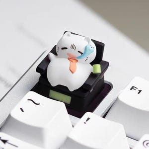 May include: A white cat figurine wearing an orange tie and a blue bandage, seated in a miniature black chair on a keyboard. The cat is holding a green cup. The keyboard keys are white.