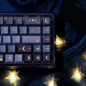May include: A custom mechanical keyboard with dark blue and purple keycaps. The keycaps feature celestial designs like moons, stars, and suns. The keyboard has a dark blue frame and the text "All you think is star" is visible on one of the keys.