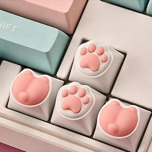 May include: Close-up of a keyboard with custom keycaps. The keycaps are white with pink paw prints and pink cat butt designs. Other keys are pastel pink and blue. The keyboard is a light color.