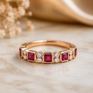 May include: A rose gold band ring featuring a repeating pattern of square-cut vibrant red rubies and small round white diamonds. The gemstones are set in a bezel style along the top half of the metal band, creating a symmetrical, alternating design.