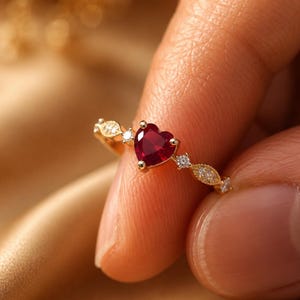 Può includere: Un anello in oro con una pietra preziosa rossa centrale a forma di cuore. La fascia è decorata con piccole pietre chiare, rotonde e a forma di marquise sui lati, creando un aspetto delicato e raffinato con una finitura metallica lucida.