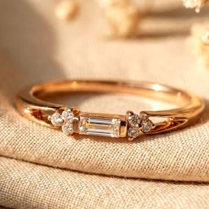 Op de afbeelding: Roségouden ring met een rechthoekige, heldere edelsteen in het midden, geflankeerd door kleinere, ronde edelstenen. De band heeft een delicaat, opengewerkt ontwerp. De ring is geplaatst tegen een neutrale stoffen achtergrond, met zachte verlichting.