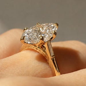 May include: A gold ring featuring a large marquise-cut center stone held by prongs, accented by a baguette-cut stone on the side. The gold band displays intricate engraved detailing along the gallery, showcasing a polished finish and reflective facets.