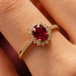 May include: A gold ring featuring a central oval-cut deep red ruby surrounded by a halo of small round white diamonds. The band is thin and also set with small white diamonds along the top half, creating a bright and elegant appearance.