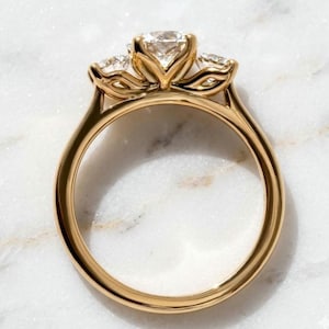 Könnte beinhalten: Ein goldener Ring mit einer Drei-Stein-Fassung, bestehend aus einem großen runden Mittelstein, der von zwei kleineren runden Steinen flankiert wird. Der Metallring ist glatt und poliert und zeigt ein klassisches Design mit Krappenfassungen.