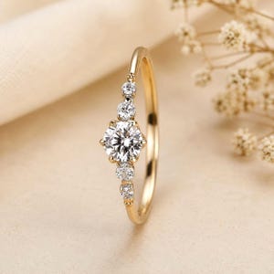 Könnte beinhalten: Ein goldener Ring mit einem großen, runden, klaren Edelstein als Mittelpunkt, flankiert von kleineren, klaren Edelsteinen. Der Ring hat ein zartes Band und ist vor einem weichen, neutralen Hintergrund platziert. Der Ring ist ein Schmuckstück.
