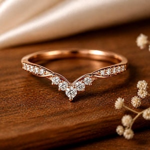 Pode incluir: Anel de ouro rosa com um design curvo, cravejado com vários diamantes pequenos. O anel tem uma aparência delicada e elegante, perfeito para uma ocasião especial. O anel é exibido em uma superfície de madeira.