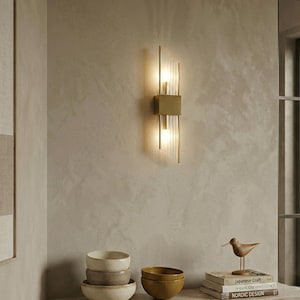以下が含まれることがあります： 長方形の真鍮ベースと2つの垂直ライトを備えたモダンなウォールスコンス。各器具は、暖かく魅力的な輝きを放つ透明なガラス管のデザインです。テクスチャ加工されたニュートラルカラーの壁に取り付けられています。