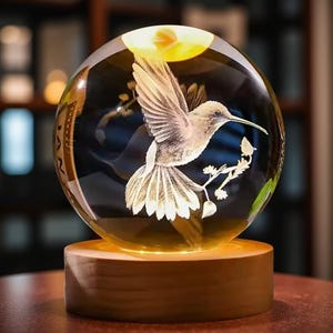 Puede incluir: Una esfera de vidrio transparente con un diseño detallado de colibrí, iluminada desde abajo por una luz cálida. La esfera se asienta sobre una base redonda de madera, creando una exhibición decorativa y artística.