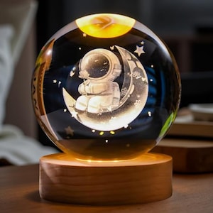 May include: A clear glass sphere with an etched design of an astronaut seated on a crescent moon, holding a flower. The sphere sits on a wooden base that emits a warm glow. Stars are etched around the astronaut and moon.
