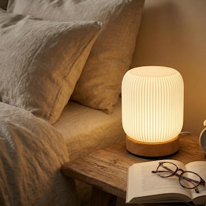 Puede incluir: Una lámpara de noche blanca con diseño acanalado se encuentra sobre una mesita de noche de madera. La lámpara tiene una base de madera y está junto a un libro abierto y gafas. Ropa de cama suave en el fondo.