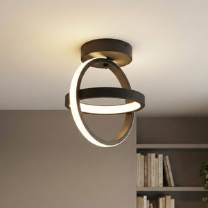 May include: Modern black ceiling light fixture with two intersecting circular rings. The inner rings emit a soft, warm white light. The fixture is mounted on a dark ceiling, with a bookshelf visible in the background.