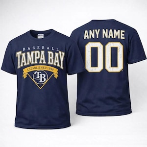 Puede incluir: Camiseta de béisbol azul marino con texto "TAMPA BAY" en blanco y dorado, establecida en 1998. La parte trasera de la camiseta muestra "ANY NAME" sobre el número "00" en blanco y dorado.