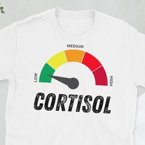 Peut inclure: T-shirt blanc avec un graphique de jauge indiquant le taux de cortisol. La jauge varie du vert au rouge, indiquant des niveaux bas à élevés. Le mot "Cortisol" est imprimé en gras en dessous.