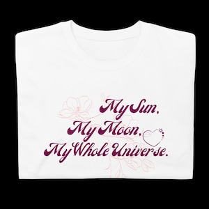 Peut inclure: T-shirt blanc avec l'inscription "My Sun, My Moon, My Whole Universe" en script violet foncé. Le motif comprend des accents floraux et en forme de cœur en violet clair.