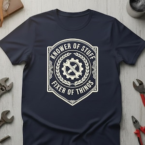Op de afbeelding: Marineblauwe t-shirt met een witte opdruk met de tekst "KNOWER OF STUFF FIXER OF THINGS". De opdruk toont een sleutel en tandwiel in een decoratief kader. Gereedschap is rondom het t-shirt zichtbaar.