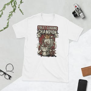Op de afbeelding: Wit T-shirt met een humoristische afbeelding. De afbeelding toont een toilet dat overstroomt met puin, een kroon en de tekst "TOILET CLOGGING CHAMPION" in rood en zwart. Het ontwerp is gecentreerd op de voorkant van het shirt.