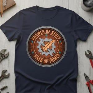 Peut inclure: T-shirt bleu marine avec un motif circulaire. Le motif comprend une clé et un engrenage, avec une plume superposée. Le texte indique "KNOWER OF STUFF" et "FIXER OF THINGS". Le design est en marron, orange et gris.