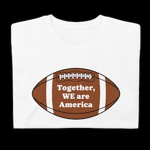 Può includere: T-shirt bianca con grafica di pallone da football americano marrone, strisce bianche e la scritta "Together, WE are America". La maglietta è realizzata in un materiale morbido e confortevole.