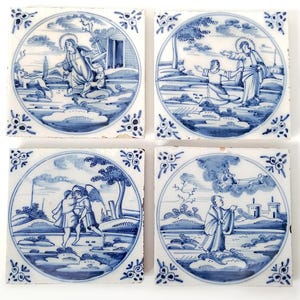 May include: Four square ceramic tiles featuring blue and white hand-painted illustrations of biblical scenes. Each tile displays a circular central motif with detailed landscape backgrounds, figures in robes, and decorative corner floral patterns.