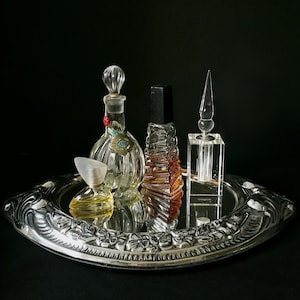 May include: A collection of perfume bottles on a silver tray. The bottles vary in shape and size, with clear glass and amber hues. The tray has a decorative floral border, set against a black background.