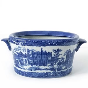 May include: A blue and white ceramic planter with a classic Willow pattern design. The planter has a rectangular shape with rounded corners, two handles, and a blue rim. The detailed pattern depicts a landscape scene with buildings and figures.