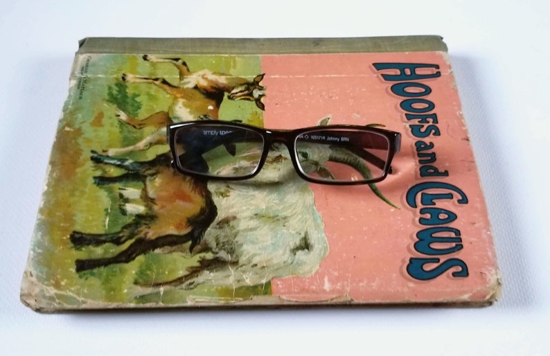 May include: A vintage children's book titled "Hoofs and Claws" with a pair of brown eyeglasses resting on top. The book cover features a colorful illustration of two goats.