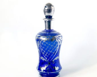 May include: A cobalt blue glass decanter with a silver-toned stopper and intricate silver detailing. The decanter has a wide base and a narrow neck.