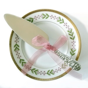 May include: A silver-plated cake server with an ornate floral embossed handle, tied with a sheer dusty pink ribbon, resting on a white porcelain plate featuring a gold rim and a delicate green leaf and purple berry pattern around the edge.