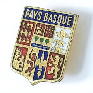 May include: A gold-toned metal pin with a shield design. The pin features the text "PAYS BASQUE" in blue, and a colorful emblem with red, green, blue, and white details. The pin is likely a souvenir or collectible item.