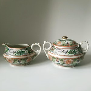 May include: A vintage porcelain tea set featuring a creamer and a lidded sugar bowl. The set is decorated with a colorful floral and bird pattern in green, orange, and gold, with a gold knob on the sugar bowl lid.