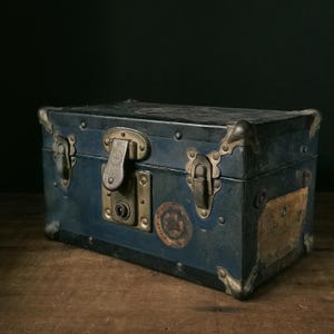 May include: A vintage blue trunk with brass hardware, corner protectors, and a central lock. The trunk has a weathered appearance, with visible wear and tear on the surface. It features decorative stickers and a rectangular label.