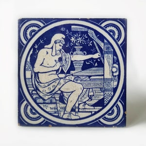 May include: Blue and white ceramic tile with a decorative design featuring a figure seated at a table with a quill and inkwell. The tile has a circular border with a floral motif and the text "The Weaver".